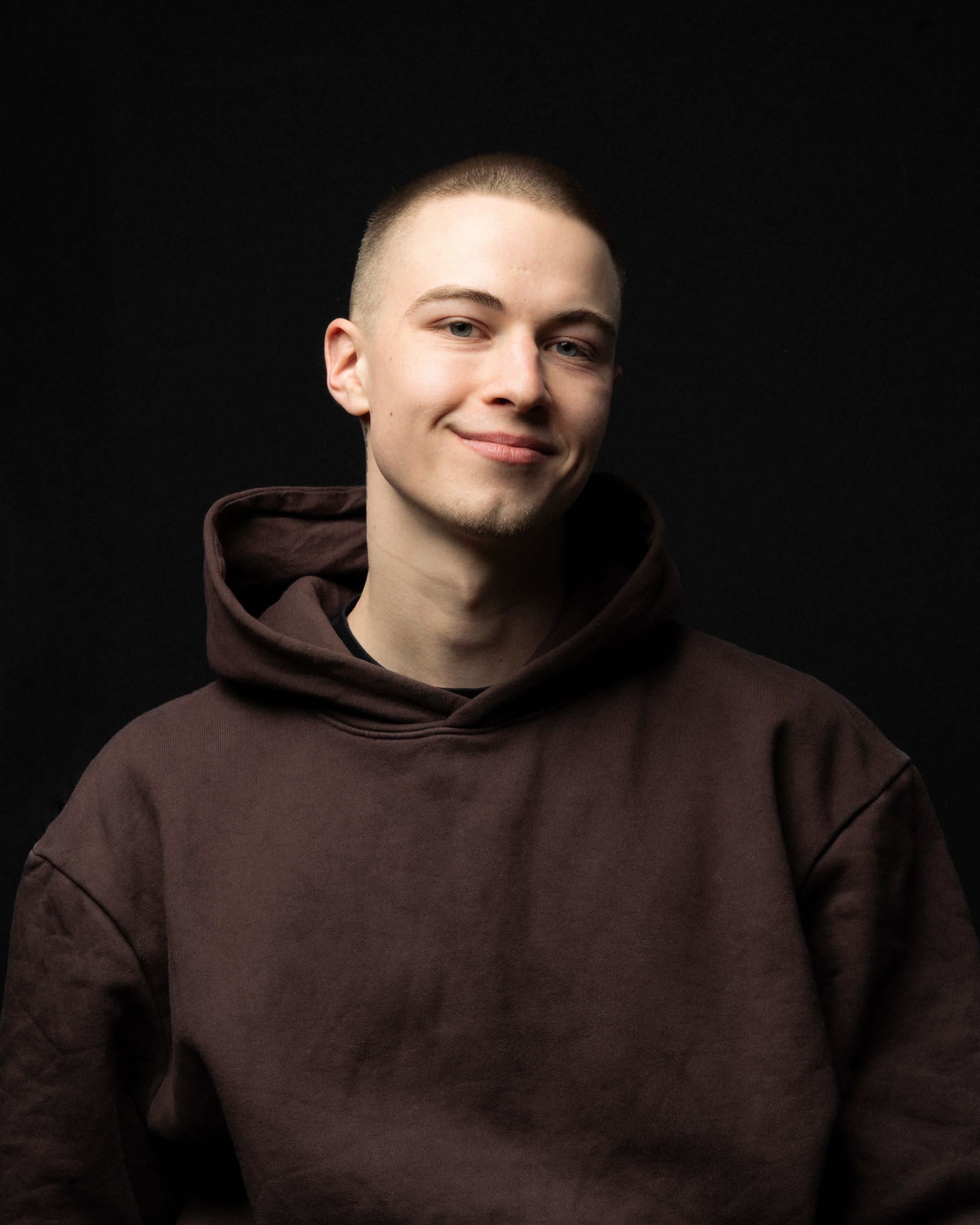 Portrait of a young man smiling in a dark hoodie against a black background.