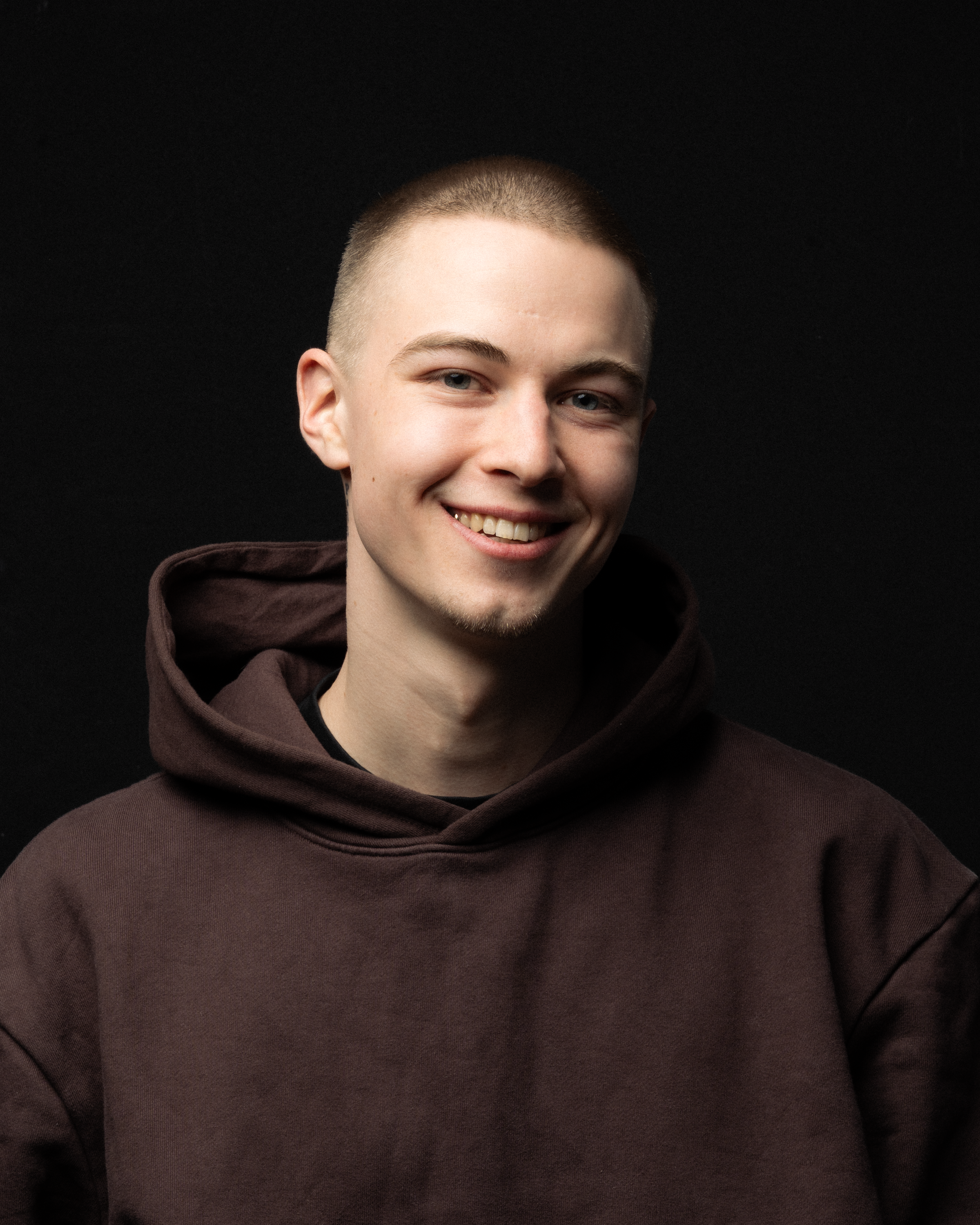 Smiling young adult in a dark hoodie against a black background.