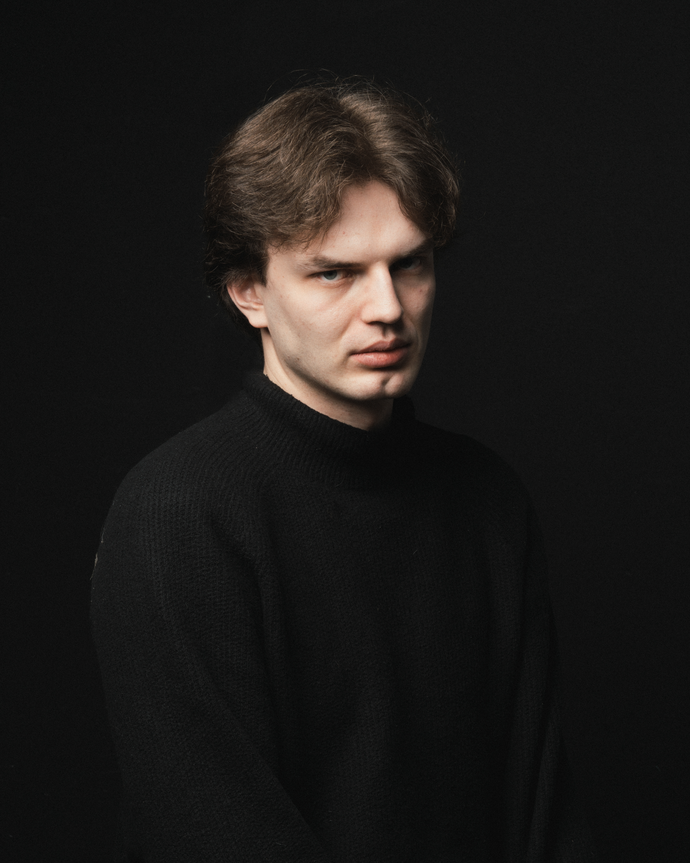 A dramatic portrait of a young Caucasian adult wearing a black sweater against a dark background.