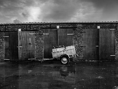 Black and white image of a trailer parked in front of old brick garages under a moody sky.