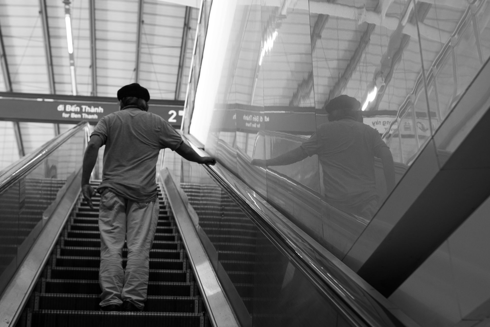 Black and white photo of a man on an escalator in a modern train station setting.