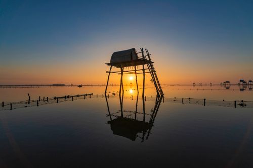 Free Stunning sunset view of a traditional Vietnamese fishing hut over calm water, reflecting vibrant colors. Stock Photo