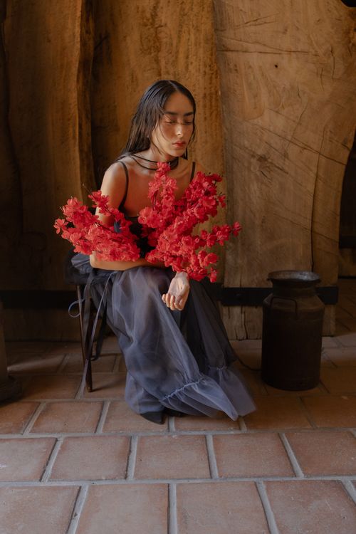 Free A woman in a dark dress holds vibrant red flowers, sitting in a rustic indoor setting. Stock Photo