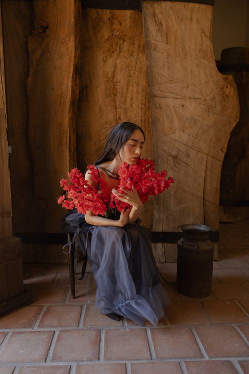 Free A woman in a flowing dress holds vibrant red flowers, seated indoors against wooden backdrop. Stock Photo
