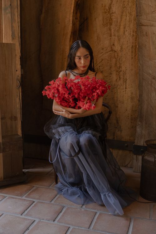 Free Woman sitting indoors in elegant dark dress holding a bouquet of striking red flowers, creating a dramatic mood. Stock Photo