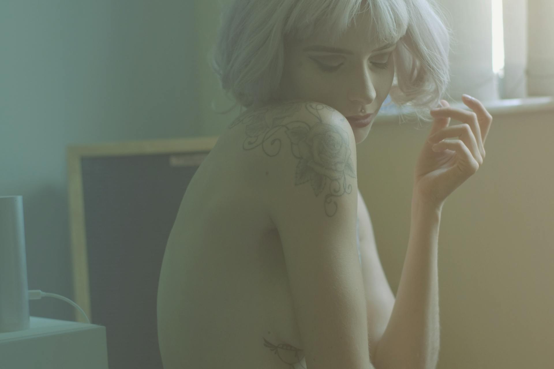 Soft-focus portrait of a tattooed woman with platinum hair, captured indoors with gentle lighting.