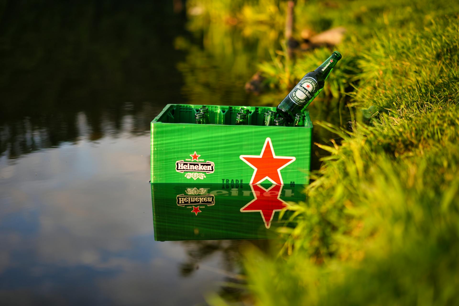 Green Heineken beer box with bottles by a calm lake, reflecting sunlight.
