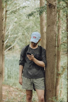 Young man in outdoor gear using smartphone while hiking in lush forest setting.