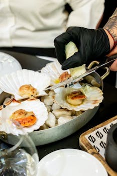 Chef adding sauce to gourmet scallops served on shells in a stylish restaurant setting.