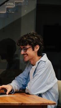 A young man wearing glasses smiles while working on his laptop in an office setting.