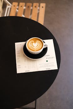 Top view of a latte with heart-shaped art served on a black table alongside a cafe menu.