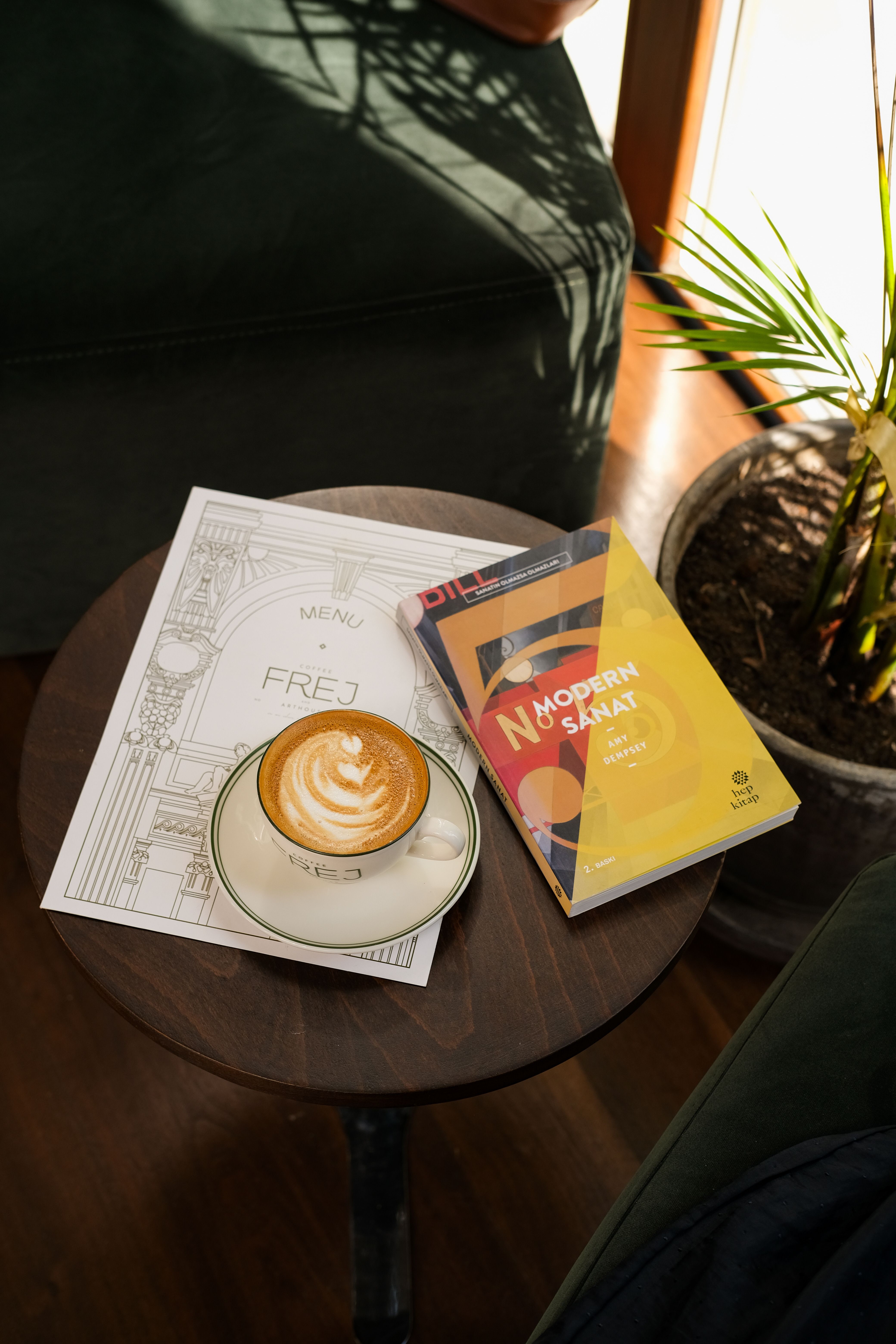 Cup of latte with intricate art on a coffee table beside a menu and book in a cozy cafe.