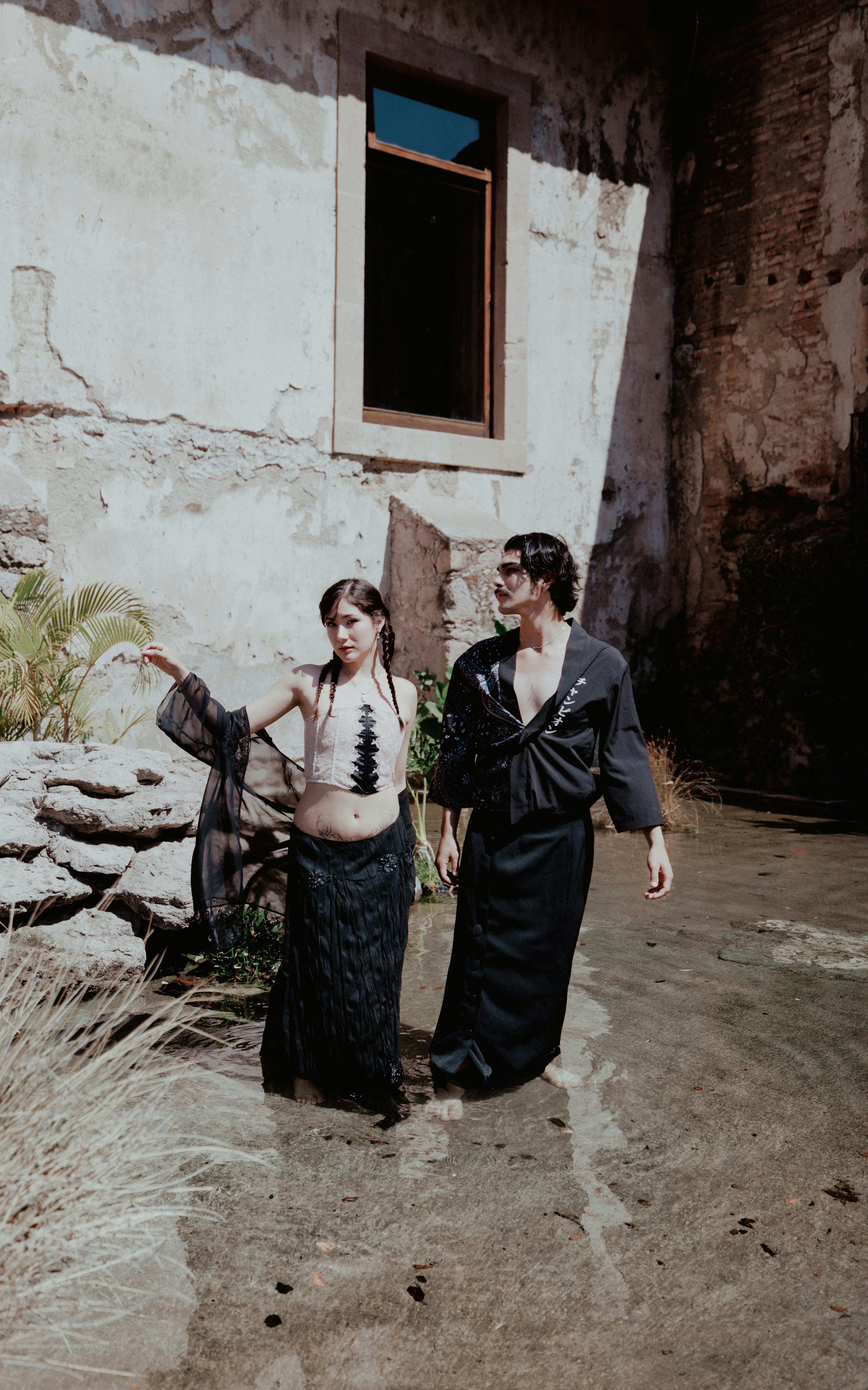 Free Stylish couple in black outfits standing in a rustic outdoor scene with water and stone walls. Stock Photo