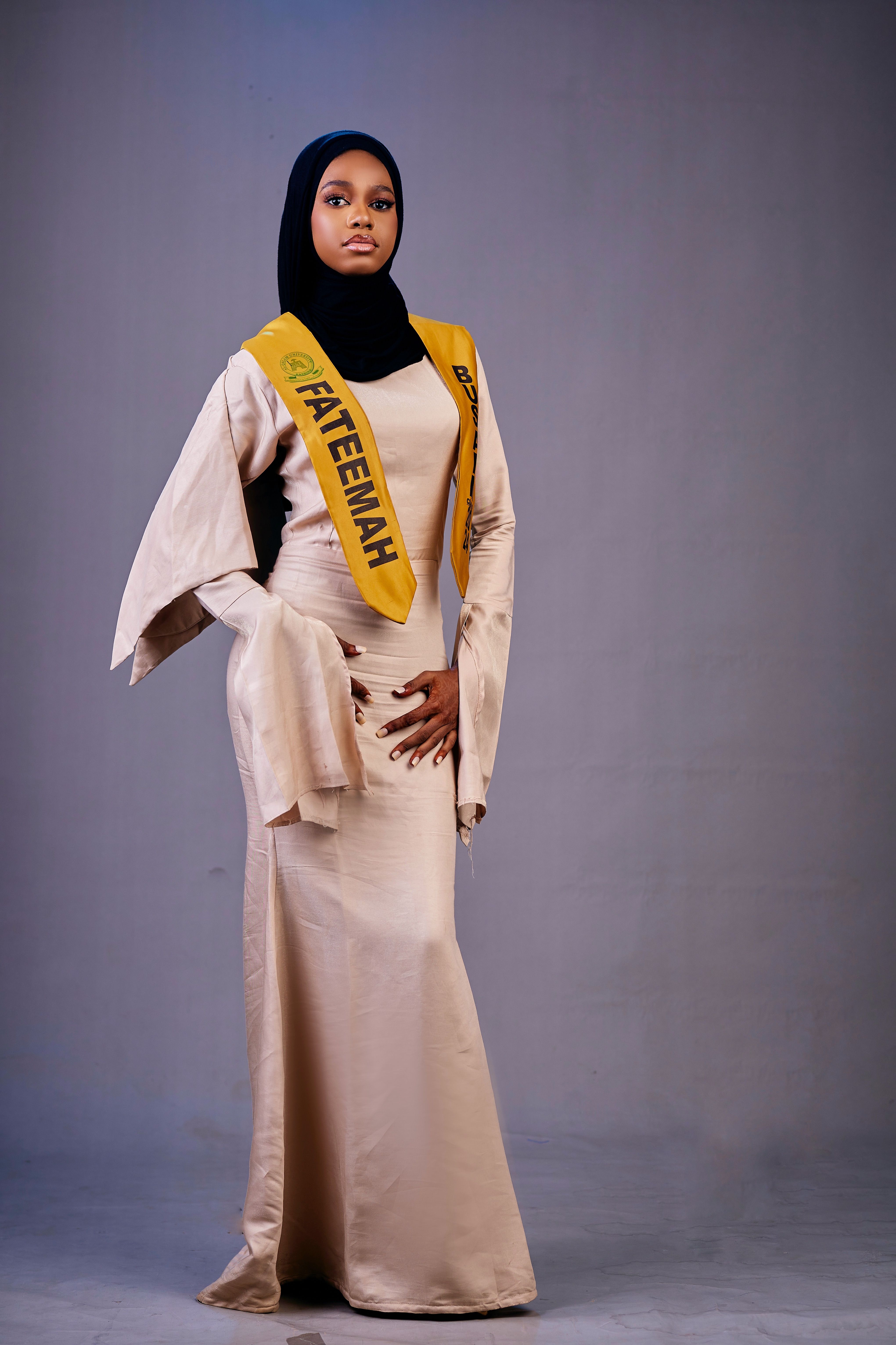 A confident woman poses in a long, elegant dress with a hijab and sash, exuding grace and style.