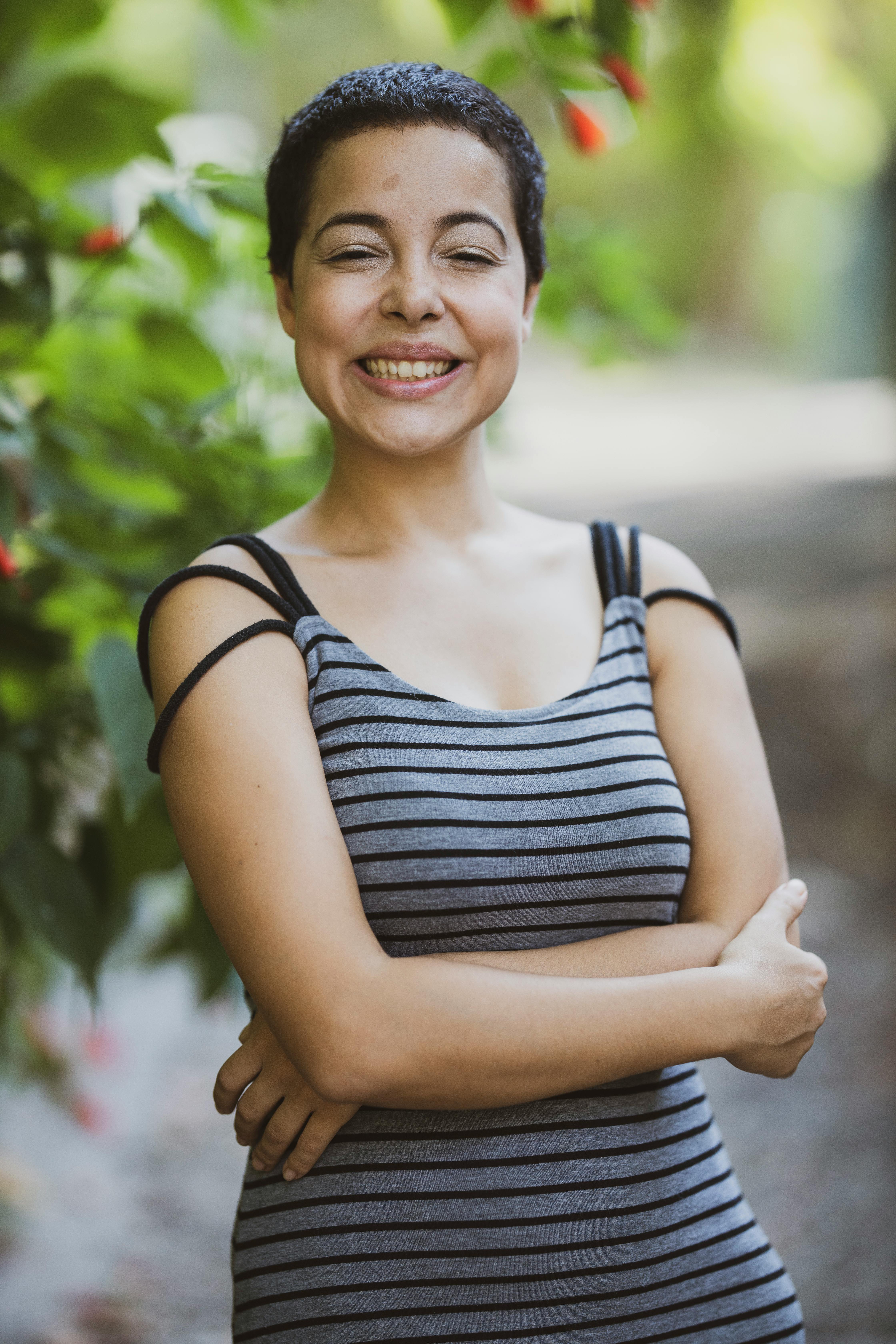 A joyful woman with short hair smiling in a striped dress, surrounded by greenery.