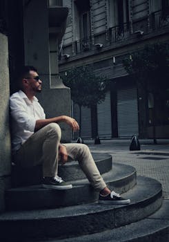 Casually dressed man in urban setting, sitting on stone steps, showcasing city lifestyle.