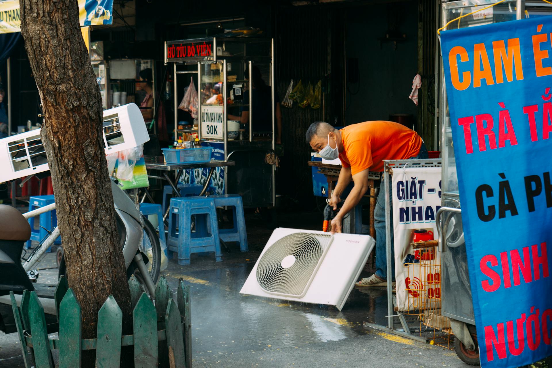 A man cleans a fan on a vibrant urban street with market stalls.