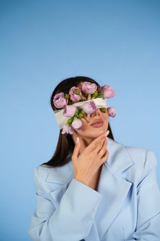 Fashionable woman in blue suit blindfolded with purple flowers against blue background.