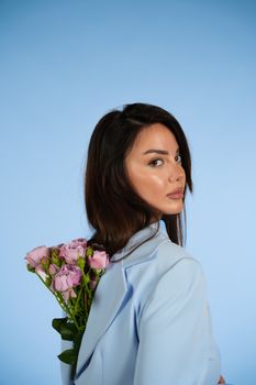 Stylish woman in pale blue suit holding pink roses against a serene blue backdrop.