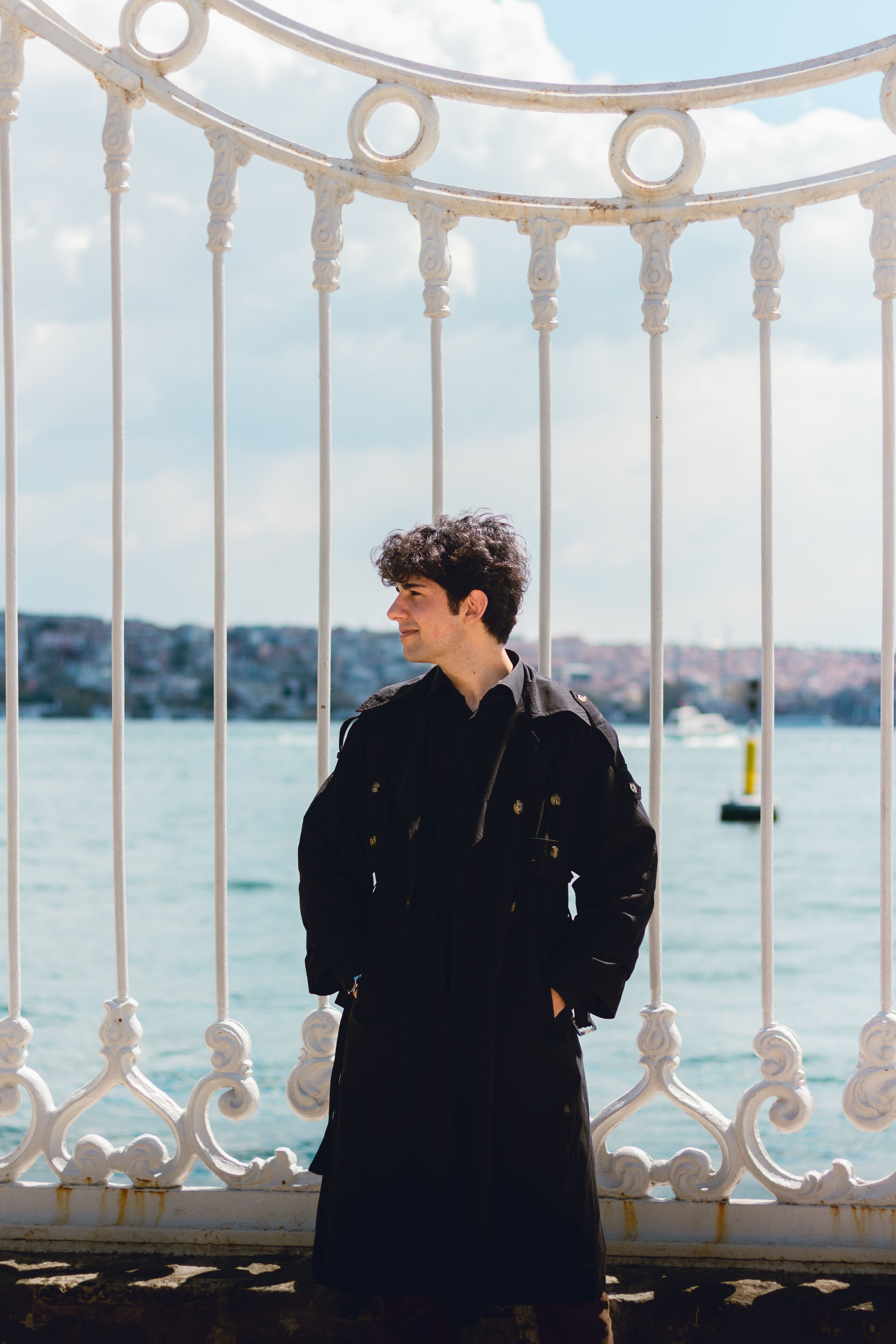 Man in black coat standing near wrought iron fence by water with city in background.