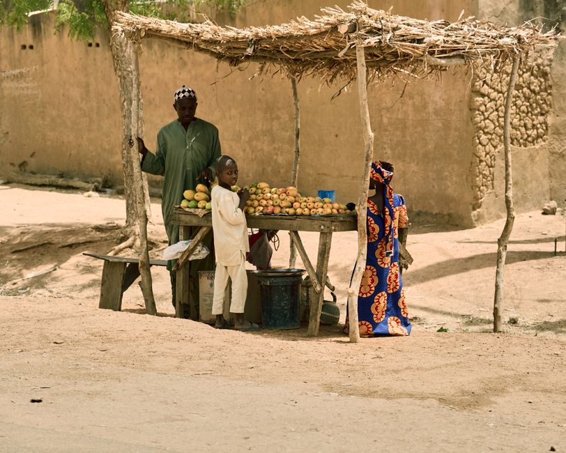 Black Nigerian family of four — mother in adire dress, father in casual shirt, two children — unloading bags of rice, beans, and vegetable oil onto a kitchen counter in a modest Lagos apartment, smiling and cheerful, bright indoor lighting, photorealistic
