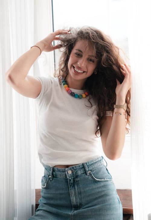 Free Smiling young woman with curly hair and colorful necklace enjoying indoors lighting. Stock Photo