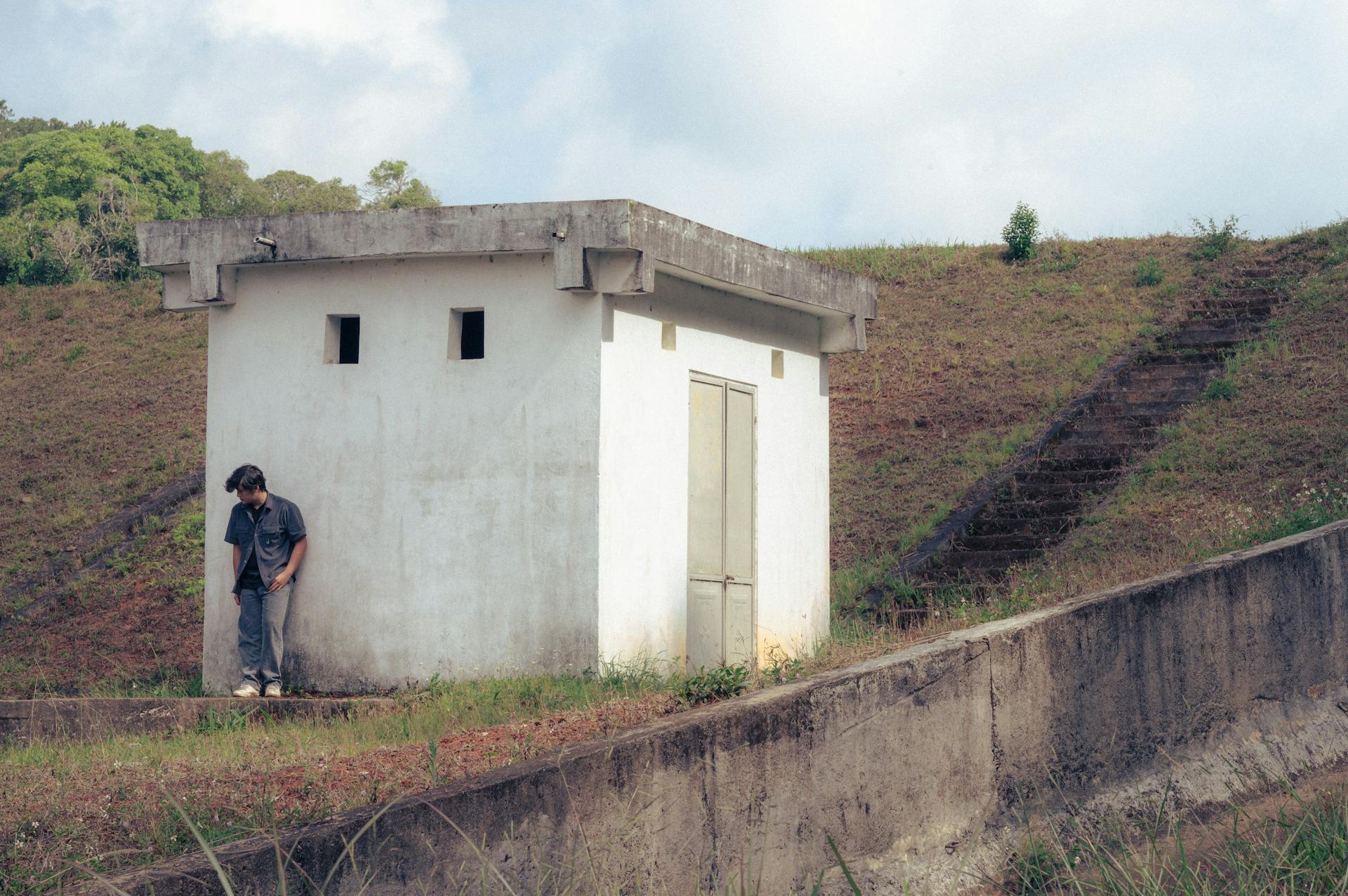 Man standing near a small utility building in a rural outdoor setting.