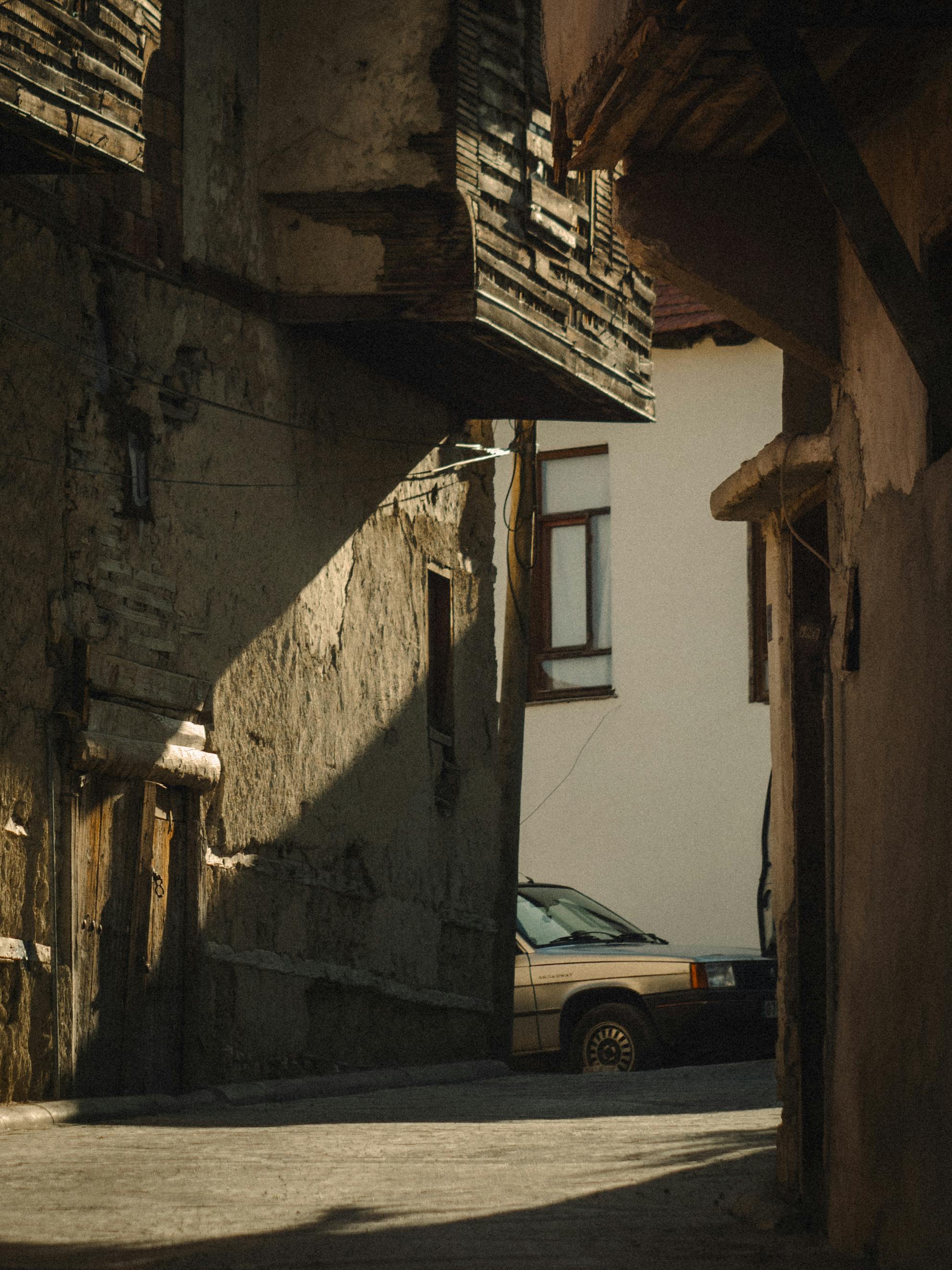 Gratuit O alee de epocă cu arhitectură rustică și o mașină veche care aruncă umbre, surprinsă în lumina caldă a zilei. Fotografie de stoc