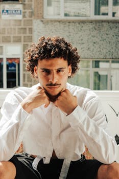 Portrait of a confident young man with curly hair wearing a white shirt, sitting indoors.
