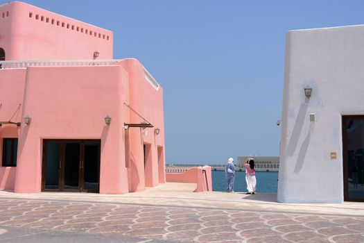 Two people walk beside pink and white Mediterranean-style buildings near the sea on a sunny day.