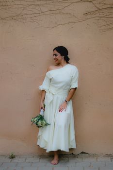 Woman in an elegant off-white dress holding a bouquet, standing against a textured wall.