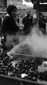 Black and white photo of a bustling street vendor with steaming chestnuts and diverse people.