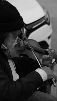 Black and white photo of a street musician playing violin next to a car.