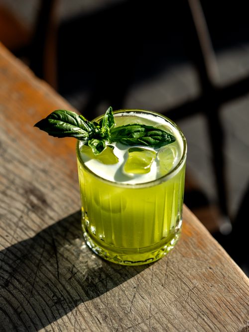 Free A vibrant green cocktail garnished with fresh basil leaves, illuminated by sunlight on a wooden table. Stock Photo