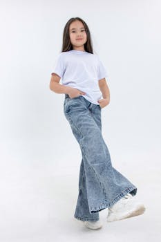 A confident young girl poses in wide-legged jeans and a white tee against a white studio background.
