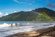 Vibrant rainbow stretches over a lush green hill by a sandy beach in Trinidad and Tobago.