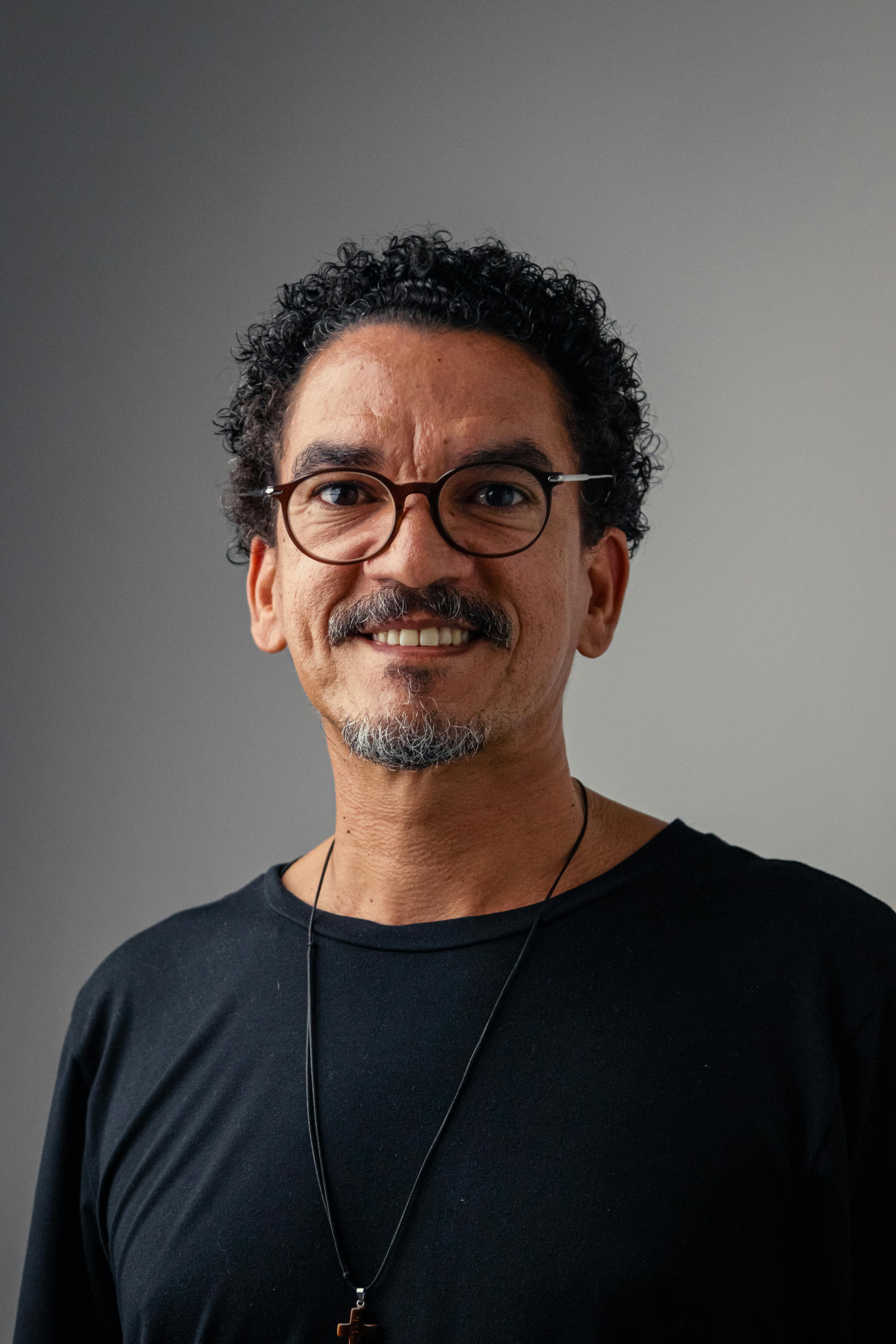 Portrait of a smiling man with curly hair and glasses against a neutral background.