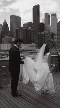 A couple in wedding attire posing on the Brooklyn Bridge with the New York City skyline in view.