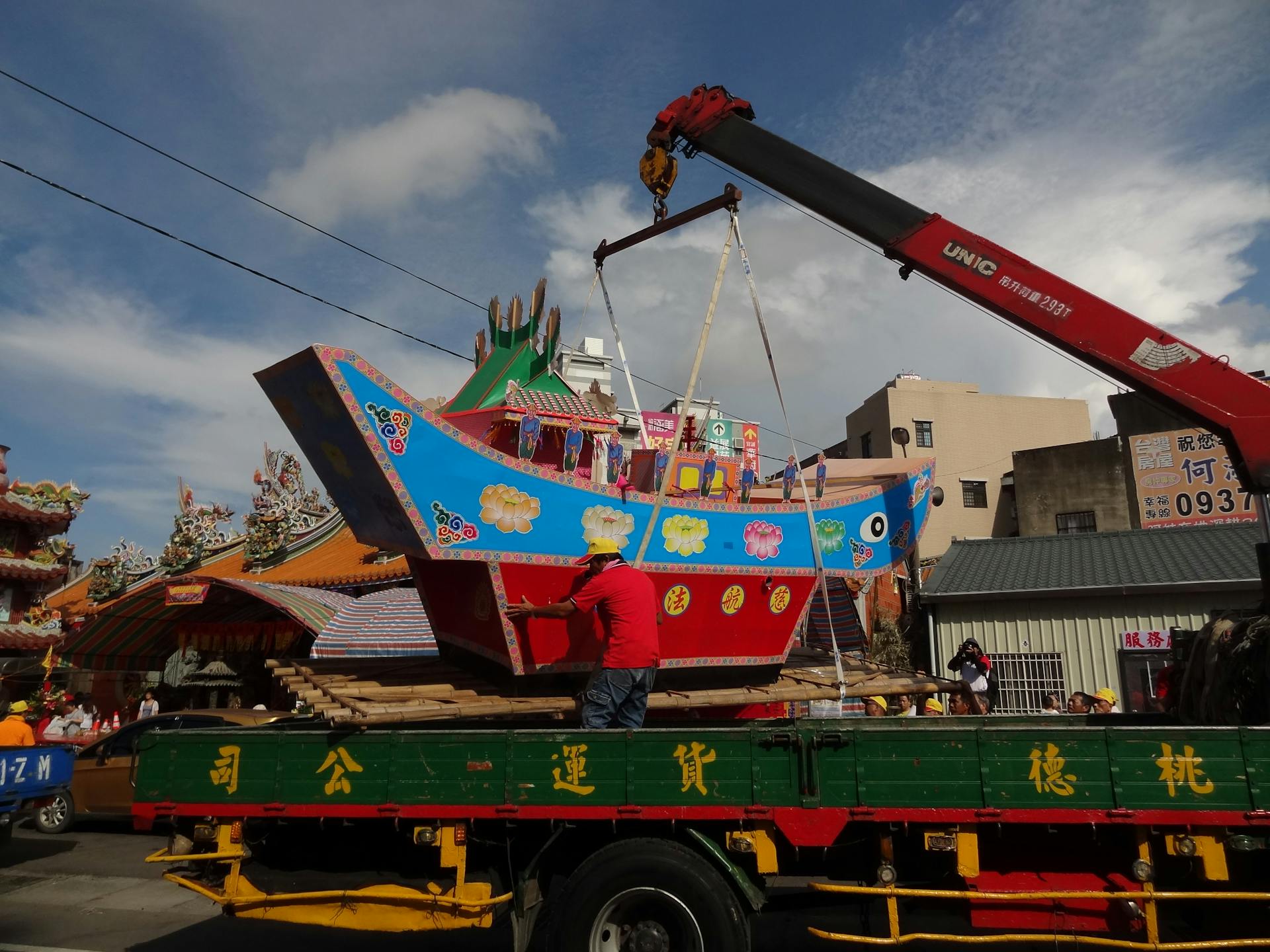 Vibrant boat being lifted by crane in a colorful outdoor festival setting.