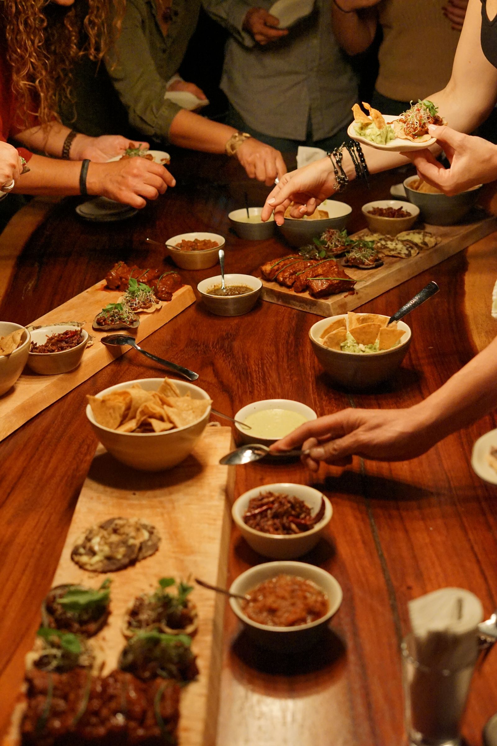 Many hands reaching across a long wooden table of small bowls and sharing plates