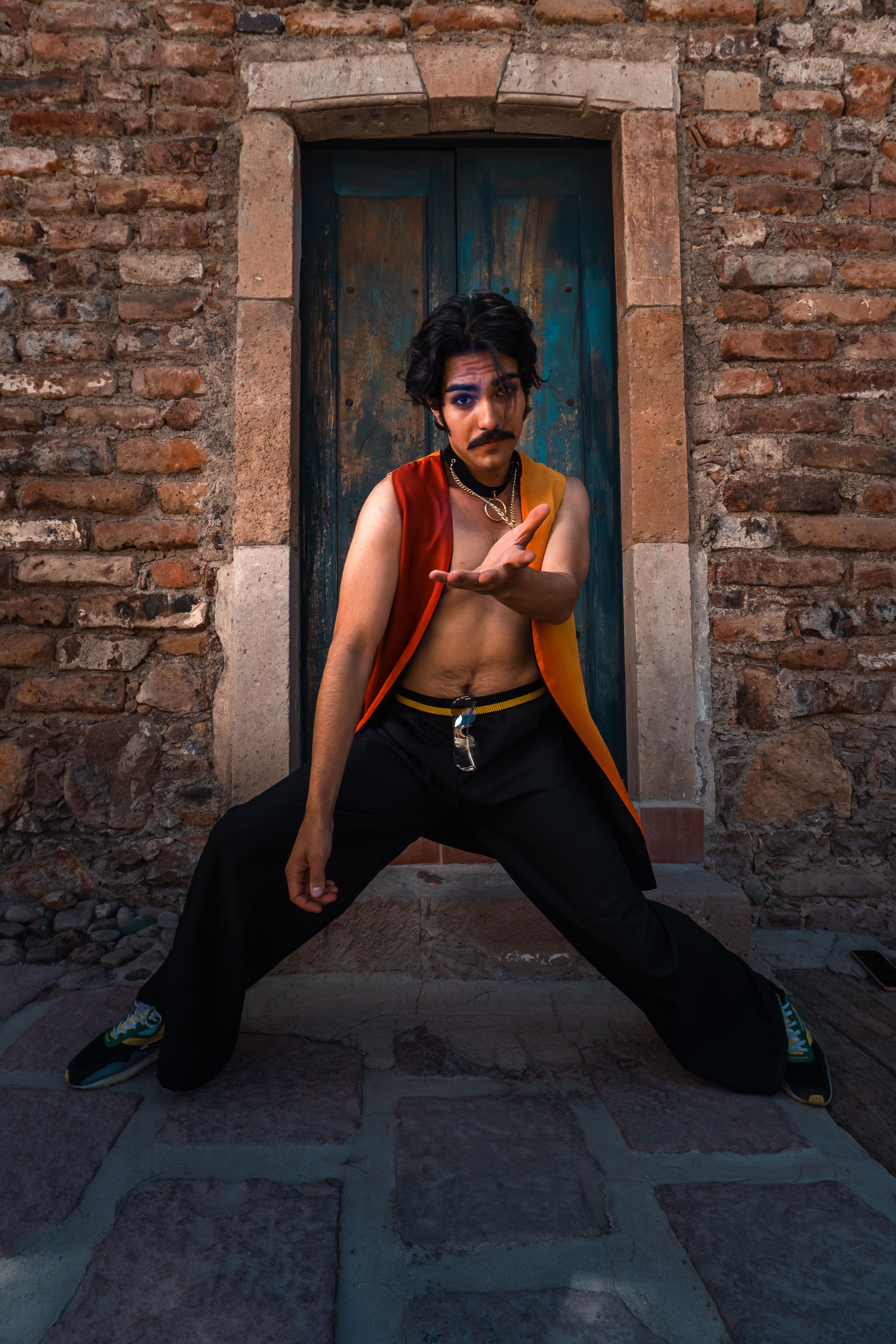 A fashionable man in a colorful outfit strikes a unique pose against a rustic brick wall.
