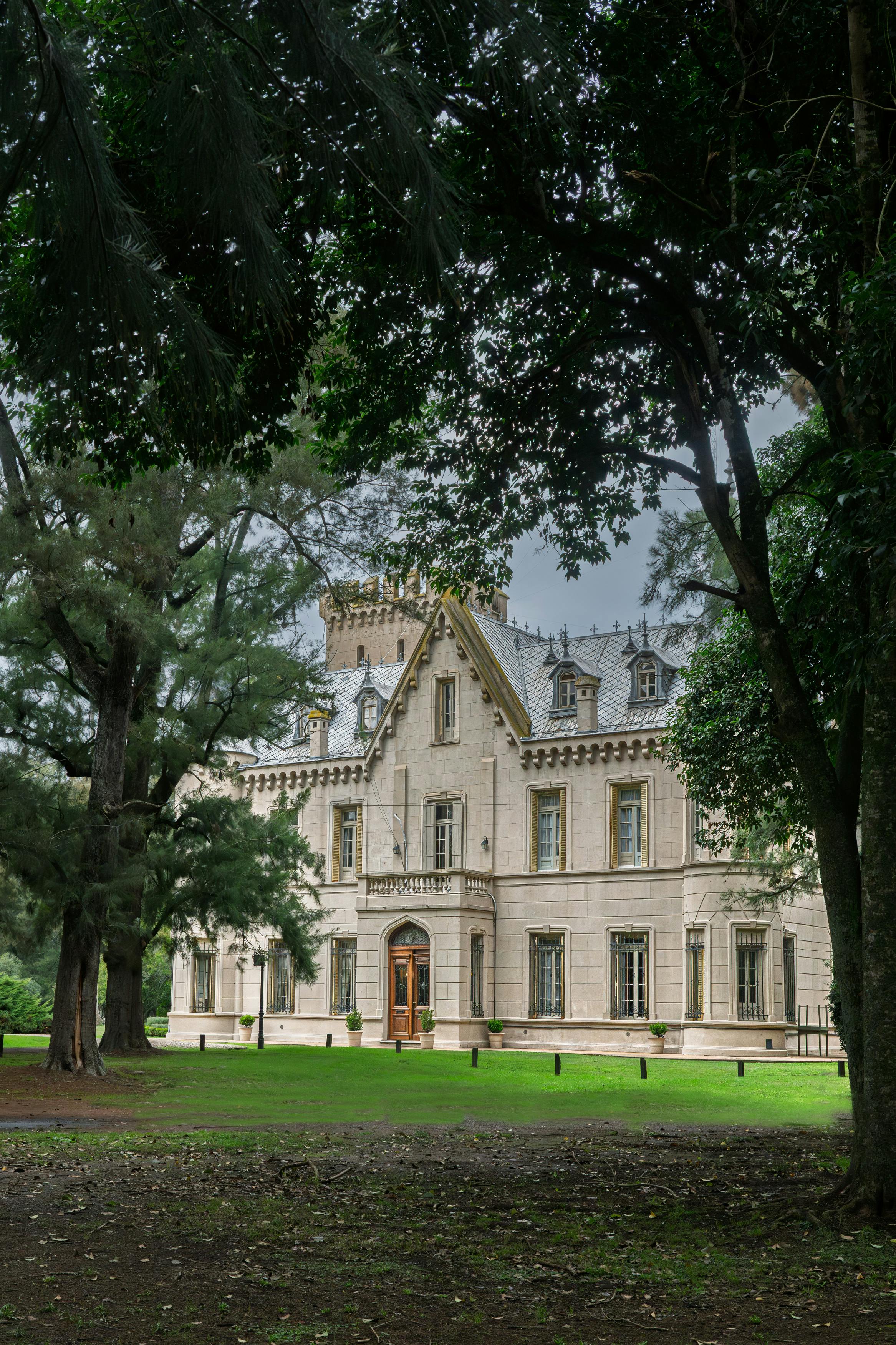 Elegant historic mansion in Lobos, Argentina, framed by towering trees and vibrant greenery.
