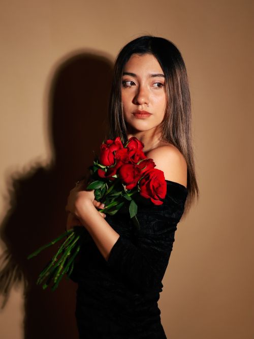 Free A young woman in a black dress holds a bouquet of red roses, posing against a simple background. Stock Photo
