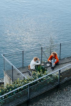 Two adults having a conversation by the water, enjoying a peaceful moment with drinks outdoors.