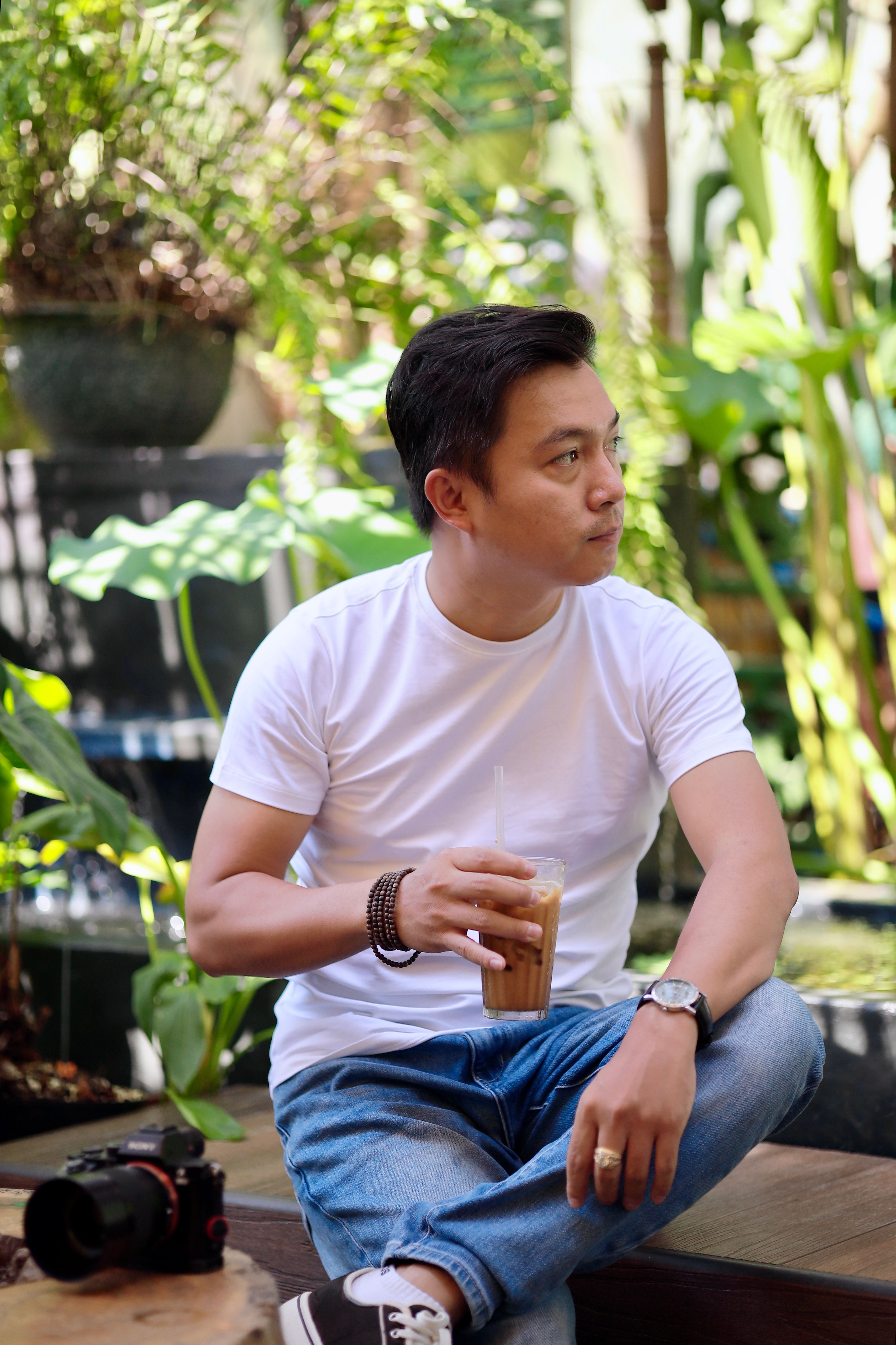 Casually dressed man drinks iced coffee in a lush outdoor garden, camera beside him.
