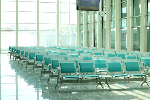 Bright airport waiting area featuring rows of empty turquoise chairs and large windows.