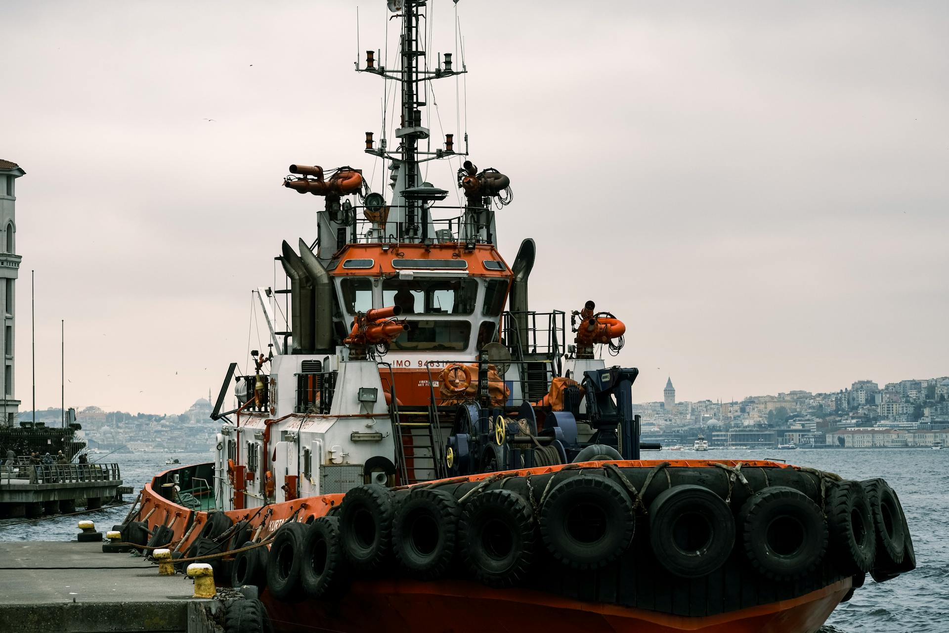 A tugboat moored at a harbor with a cityscape backdrop on a cloudy day.