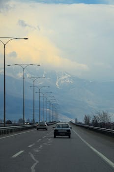 Cars drive along a highway with street lamps and a mountain backdrop under cloudy skies.