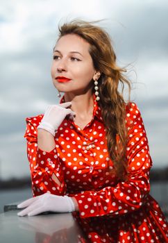 Stylish woman in a red polka dot dress and white gloves posing outdoors against a cloudy sky.