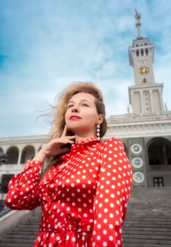 Stylish woman in red polka dot dress poses in front of a historic tower on a bright day.